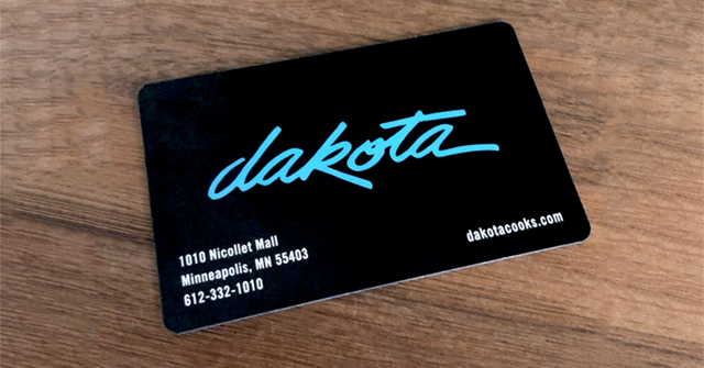 A black Dakota card with blue script text, displaying contact information for Dakota in Minneapolis, MN, on a wooden surface.